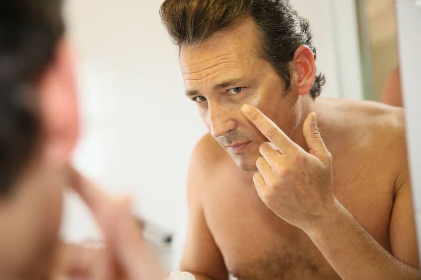 MIddle age man in front of a mirror and applying cream to his under eyes